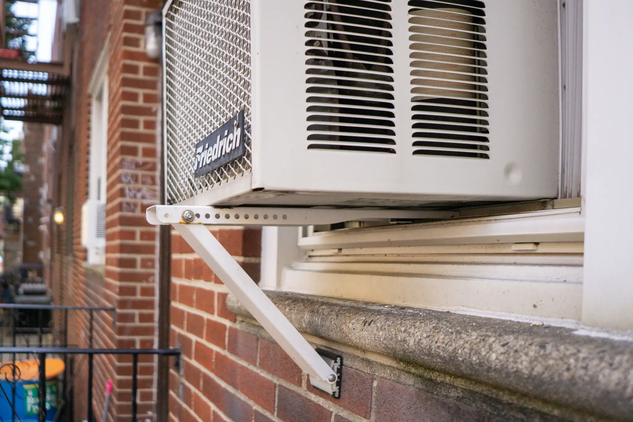 A window air conditioner installed in a double-hung window with a manufacturer-approved exterior support bracket underneath, viewed from indoors in a real home