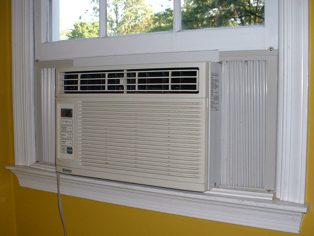 An indoor view of a window air conditioner in a double-hung window showing the gap above the unit and the lower sash lowered down to a filler panel area